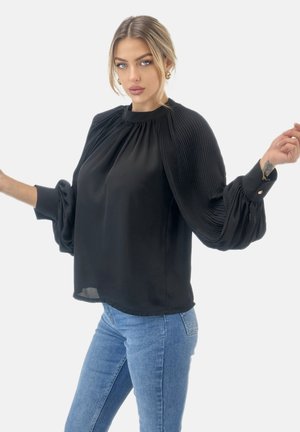 Blusa negra con mangas plisadas, escote redondeado y puños abotonados. Tejido suave y liso con un corte relajado. Combinada con vaqueros azul claro.