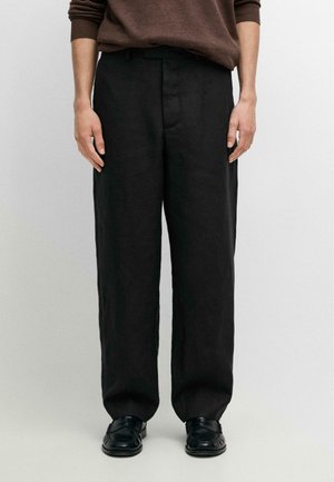 Person iført løstsiddende sorte bukser, brun langærmet top og sorte loafers, stående mod en ensartet lys baggrund.