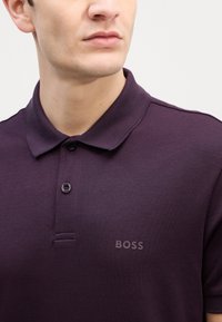 Tmavofialová polo košeľa vyrobená z hladkej tkaniny. Disponuje golierom, dvoma gombíkmi a decentným logom "BOSS" v zodpovedajúcej farbe na hrudi.
