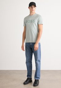 Camiseta de manga corta verde claro con el logo "BOSS", combinada con unos jeans rectos azules y zapatillas negras. El modelo lleva una gorra negra.