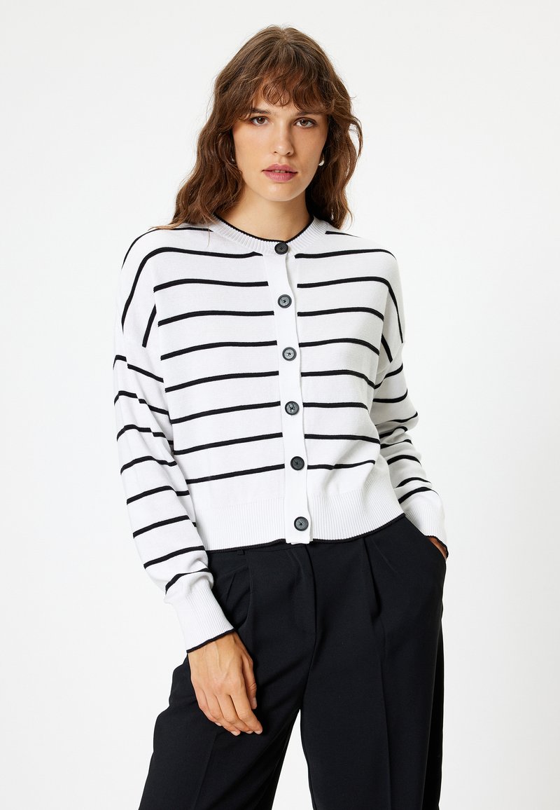 Witte cardigan met zwarte horizontale strepen, voorzien van een knoopsluiting, geribbelde manchetten en zoom. De stof lijkt zacht en lichtgewicht.