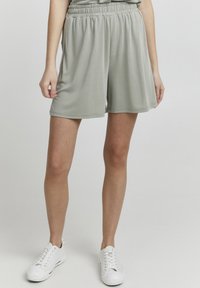 Light green athletic shorts with an elastic waistband, mid-thigh length, smooth texture, and side pockets. Paired with white sneakers.