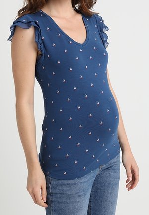 Kobieta w niebieskiej ciążowej bluzce z dekoltem w serek, ozdobionej małymi białymi i czerwonymi nadrukami żaglówek oraz falbanami przy rękawach, w połączeniu z niebieskimi dżinsami.