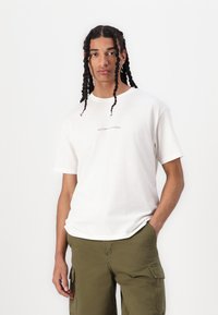 Hvid bomulds t-shirt med teksten "WELCOME TO PARADISE" i lysegrå, parret med olivengrønne cargo-shorts.