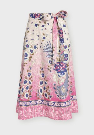 Femme debout portant un haut blanc à manches courtes, une jupe midi fleurie rose et bleue, des sandales blanches et tenant une pochette blanche.