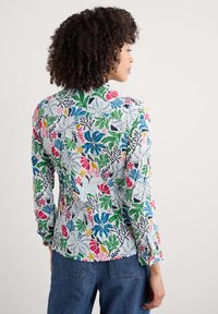 Chemise �à boutons avec un motif floral, présentant un fond bleu clair avec des accents verts, roses, jaunes et navy, fabriquée en tissu texturé.