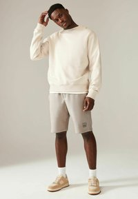 Lys beige sweatshirt med ribbede manchetter, parret med lyse grå shorts med et logo-patch. Biske sneakers og hvide sokker fuldender outfittet.
