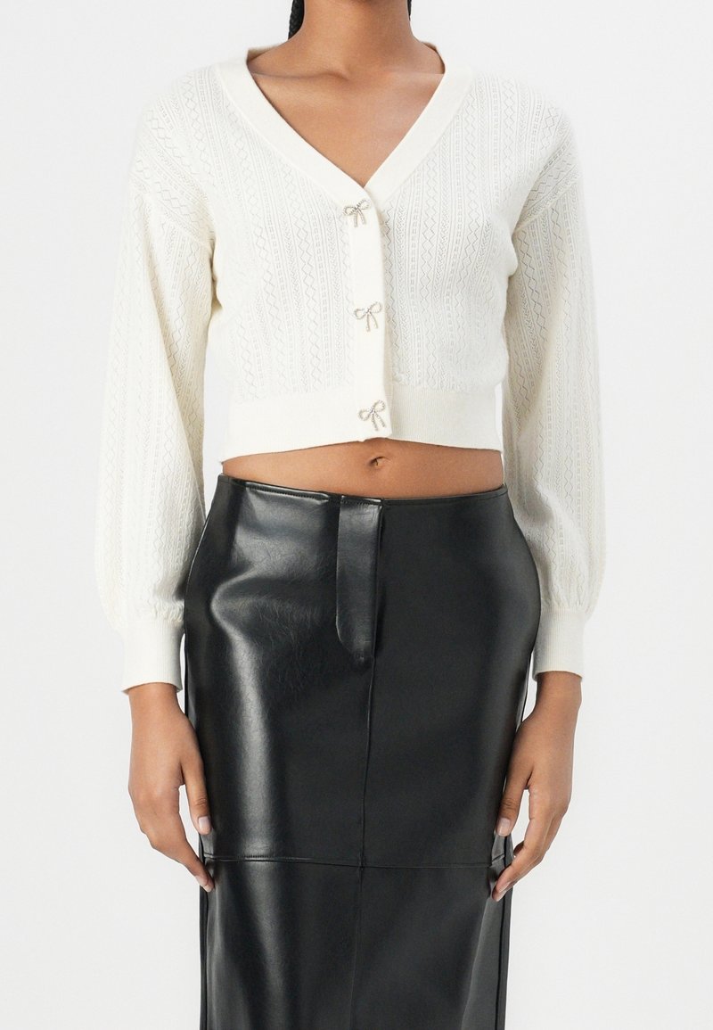 Femme portant un cardigan court en maille blanche avec des boutons en forme de nœud et une jupe en cuir noire taille haute, debout devant un fond blanc.