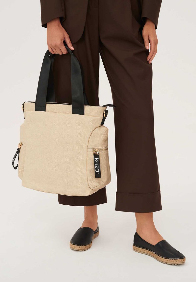 Beżowa torba wykonana z płótna z czarnymi uchwytami, z przednimi kieszeniami na zamek błyskawiczny i metką z logo. Gładka tekstura o minimalistycznym wzornictwie.