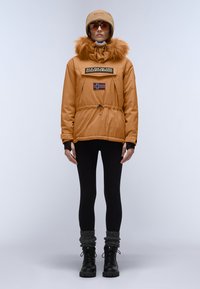 Orange pufferjakke med pelsforet hætte, prydet med et patch med et flag, sammen med sorte leggings og sorte snørebåndsstøvler.
