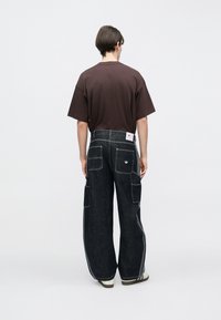 Dunkle Denim-Cargohosen mit weitem Bein, mehreren Taschen und weißen Steppnähten. Kombiniert mit einem braunen Kurzarm-T-Shirt.