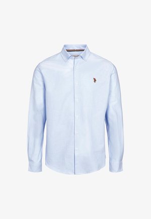 Man draagt een lichtblauw overhemd met knopen en een klein geborduurd logo, donkere spijkerbroek en een bruine leren riem met metalen gesp.
