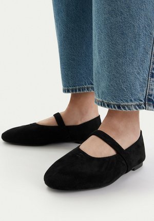 Füße in schwarzen Wildleder-Mary-Jane-Ballerinas, kombiniert mit blauen cropped Denim-Jeans vor weißem Hintergrund.