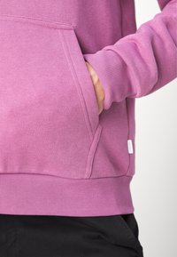 Personne portant un sweat-shirt mauve à manches longues avec la main dans la poche kangourou avant, associée à un pantalon noir sur un fond uni.