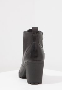 Botín de cuero negro con tacón grueso, textura suave y una asa en la parte trasera. Presenta detalles de costura y un logotipo en el lado.