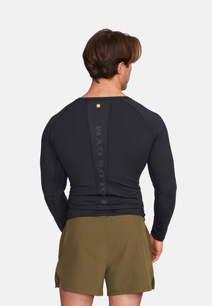 Man wearing black long-sleeve athletic shirt with vertical "Gold's Gym" text on back and olive green shorts, standing facing away.