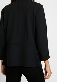 Blazer noir à coupe décontractée, doté de manches trois-quarts, d'un ourlet évasé et de détails de couture minimalistes. Texture lisse et mate.