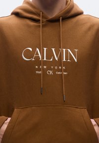 Brun bomulls-hoodie med framficka i kängurstil, huva med dragsko och vitt broderat logotyp med texten "CALVIN NEW YORK" och varumärkets initialer.