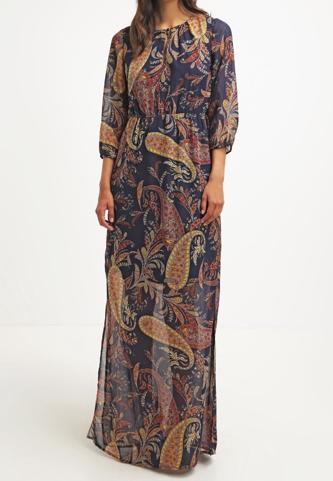 Femme portant une longue robe bleu marine avec des motifs cachemire orange, jaune et rouge et des manches trois-quarts sur fond blanc.