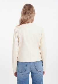 Giacca in ecopelle crema con un taglio strutturato, maniche lunghe e dettagli delle cuciture ben visibili, abbinata a jeans a vita alta.