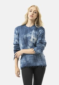 Pull en maille bleue avec motif texturé, ourlet effiloché et manches froncées. Le matériau semble doux et légèrement usé.