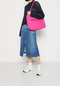 Bolso de hombro rosa con "SAMSØE" en letras negras, combinado con una blusa de punto azul marino y una falda de mezclilla con una abertura frontal. Zapatillas blancas.