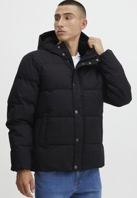Veste noire rembourrée avec capuche, dotée d'une fermeture à bouton et à zip, de poches latérales et d'une texture matelassée. Portée par-dessus une chemise blanche.