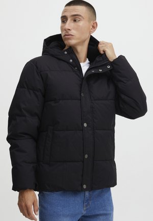 Schwarze gepolsterte Jacke mit Kapuze, ausgestattet mit einem Knopf- und Reißverschluss, Seitentaschen und einer gesteppte Textur. Getragen über einem weißen Hemd.