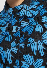 Black fabric with intricate blue floral embroidery, featuring a collar and button details, highlighting a textured lace overlay.