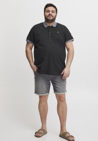 Fekete póló ing szürke gallérral és ujjal, szürke farmer sorttal párosítva. A modellen barna szandál van, fehér háttér előtt áll.
