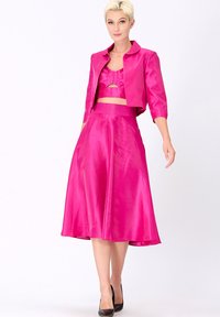 Ensemble deux pièces en satin fuchsia comprenant un haut court avec des volants et une jupe fluide, complété par une veste courte et des escarpins noirs.