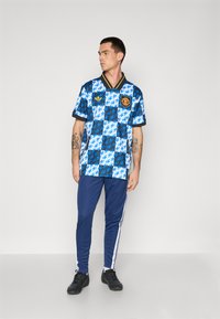 Blau-gestreiftes Fußballtrikot mit schwarzen Akzenten, gelbem Kragen und Vereinslogo, kombiniert mit navy-blauen Sporthosen und schwarzen Schuhen.