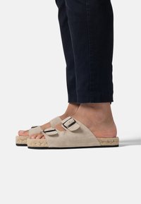 Sandales à glissière en daim beige avec deux sangles ajustables en cuir noir et boucle, semelle intermédiaire en jute tressé, et semelle extérieure en caoutchouc noir.