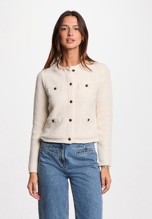 Krämfärgad kort jacka med strukturerad tyg, med rundad halsringning, fyra påsydda fickor och svarta knappar. Buren med blå denimbyxor.