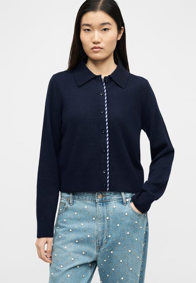 Γυναίκα που φοράει μπλε σκούρα ζακέτα με κουμπιά πάνω από ριγέ πουκάμισο και ανοιχτόχρωμα τζιν διακοσμημένα με πέρλες και μεταλλικές τρούκ, στέκεται με το ένα χέρι στην τσέπη.