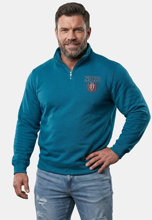 Smilende mand med kort hår og skæg iført en turkis kvartlynlåset sweatshirt med "NORD 07" og lyse revnede jeans, stående med hånd på hoften.