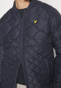 Granatowa pikowana kurtka puchowa z prążkowanym kołnierzem, zamkiem błyskawicznym i kieszeniami. Na lewej piersi znajduje się żółte logo. Gładka faktura.