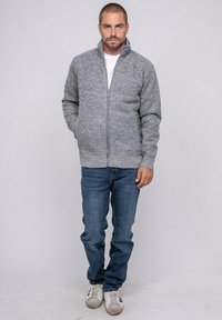 Homme debout les mains dans les poches, portant un sweat à zip gris, un jean bleu, des baskets blanches et un t-shirt blanc sur fond uni.