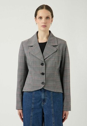 Vrouw met donker haar draagt een grijze geruite blazer met zwarte knopen, een zwart topje en hoge blauwe spijkerbroek tegen een effen lichte achtergrond.