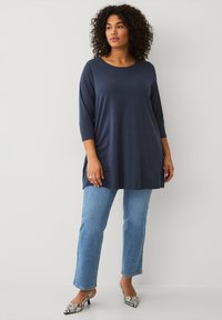 Marineblauwe tuniek met lange mouwen en een ronde hals, ontspannen pasvorm en zijsplitten, gecombineerd met lichtblauwe rechte jeans en patroonhakken.