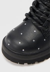 Stivaletto alla caviglia in pelle sintetica nera con borchie in cristallo, punta in pizzo testurizzato e suola in gomma spessa con trazione rigata.