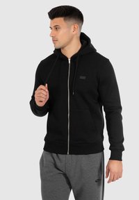 Haut à zip noir avec une poche avant, des cordons de serrage et des poignets côtelés. Porté avec un pantalon de survêtement gris. Design simple, tissu de couleur unie.