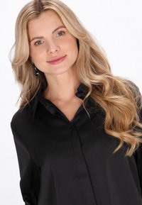 Sort satinbluse med en klassisk krave, knaplukning og en glat tekstur, båret af en model med bølget hår og øreringe med vedhæng.