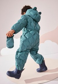 Combinaison de neige matelassée en bleu canard avec des motifs arrondis, capuche avec oreilles et poignets élastiques. L'enfant tient un gant assorti tout en portant des bottes en caoutchouc noires.