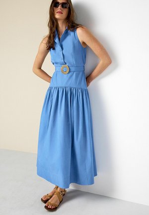 Frau trägt ein blaues ärmelloses Midikleid mit einem geflochtenen Gürtel und beigen Sandalen, steht vor einer zweifarbigen Wand in Beige und Weiß.