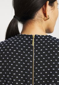 Černý látkový top s malými bílými a modrými tečkami, se viditelným zlatým zipem na zadní straně a hladkou texturou.