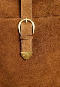 Materiale in suede marrone con una superficie liscia, caratterizzato da una fibbia in metallo dorato e un'etichetta a forma di scudo impressa con "W:11."