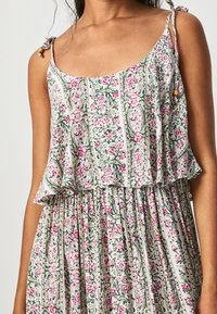 Mujer vistiendo un vestido de verano sin mangas con un estampado floral en verde, rosa y blanco, que cuenta con tirantes ajustables con cuentas de madera.