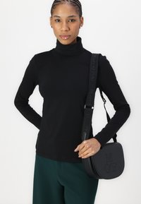Zwarte coltrui met lange mouwen, gecombineerd met groene wijde broek. Model houdt een zwarte schoudertas vast met geperste initialen "MOP."