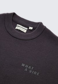 Donkergrijs crewneck sweater met ribbelkraag en zwarte vette opdruk "WHAT A VIBE" op de borst, met merk- en maatlabels binnenin de kraag.